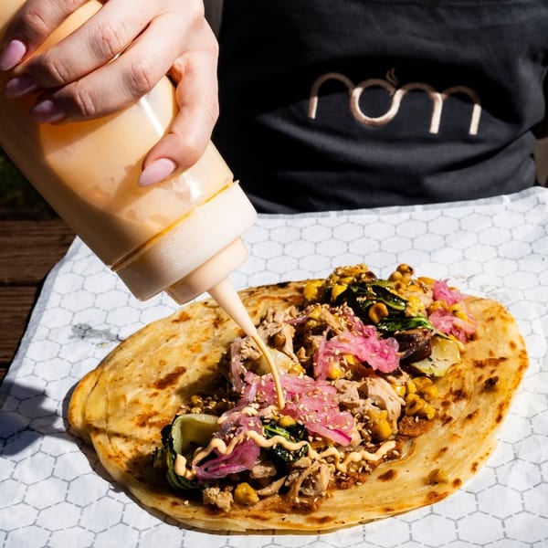 A hand squeezes sauce onto a Nom flatbread filled with meat, pickled onions, corn, and greens.