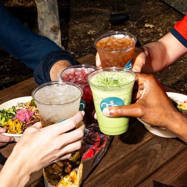 Four people toast various iced drinks and enjoy meals at an outdoor wooden table.