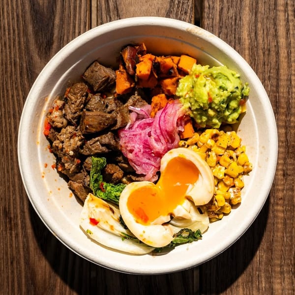Colorful bowl with beef, sweet potatoes, guacamole, corn, pickled onions, and a soft-boiled egg on wood.