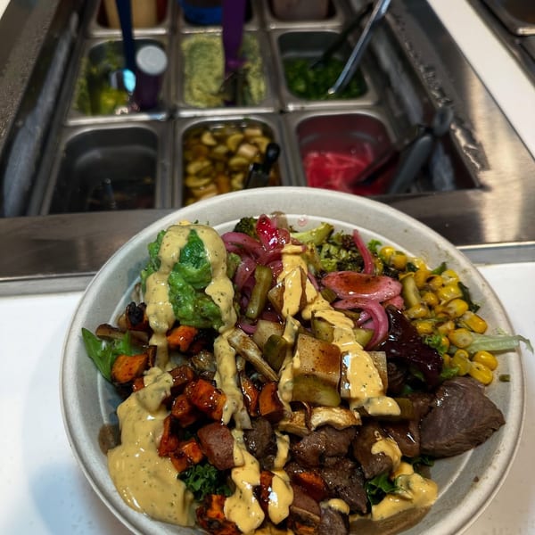 A colorful custom bowl with meat, roasted vegetables, and creamy dressing, in front of a salad bar.