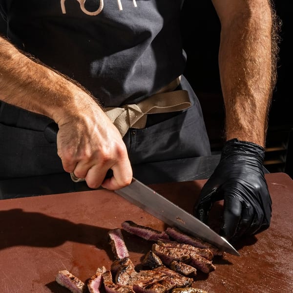 Chef slicing grilled steak on a brown cutting board, wearing an apron with the "nom" logo.