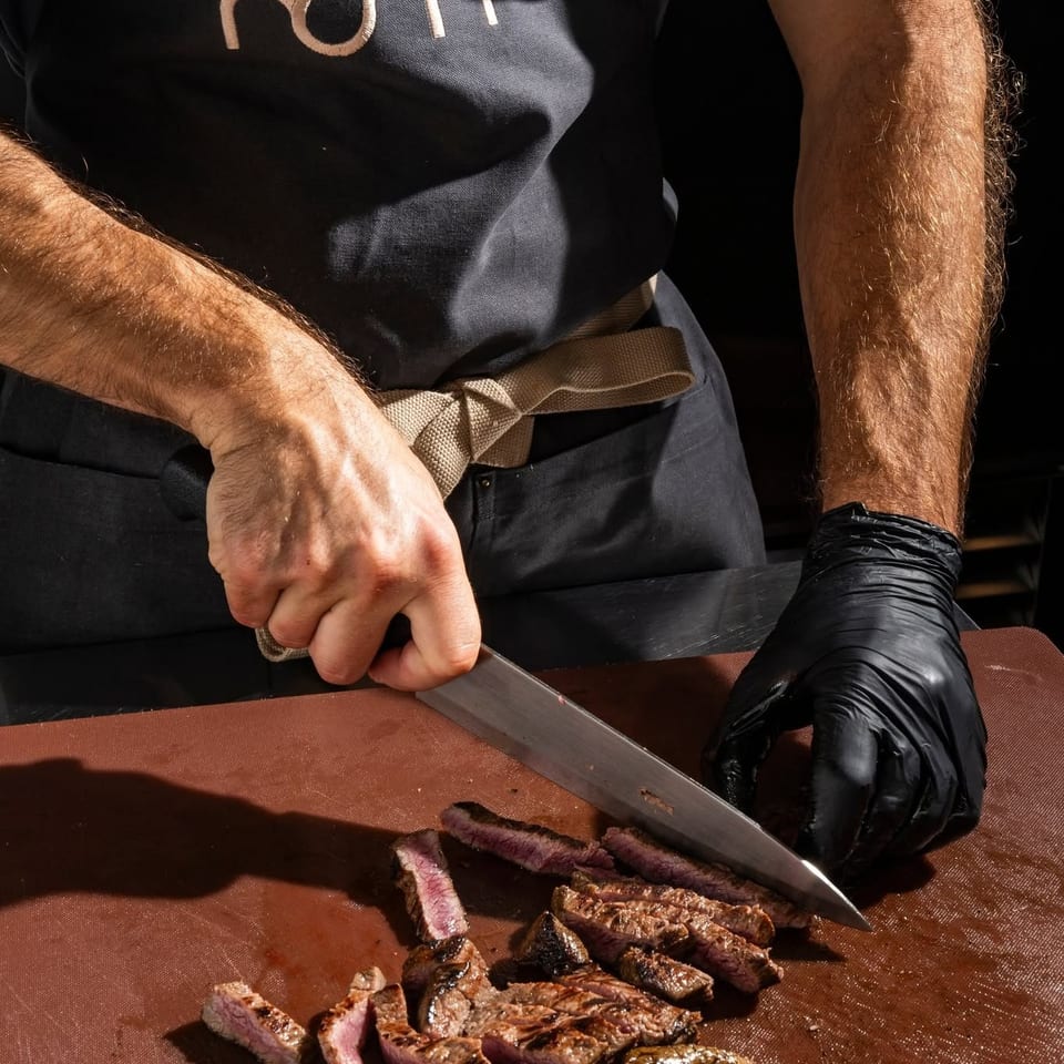 Chef slicing grilled steak on a brown cutting board, wearing an apron with the "nom" logo.