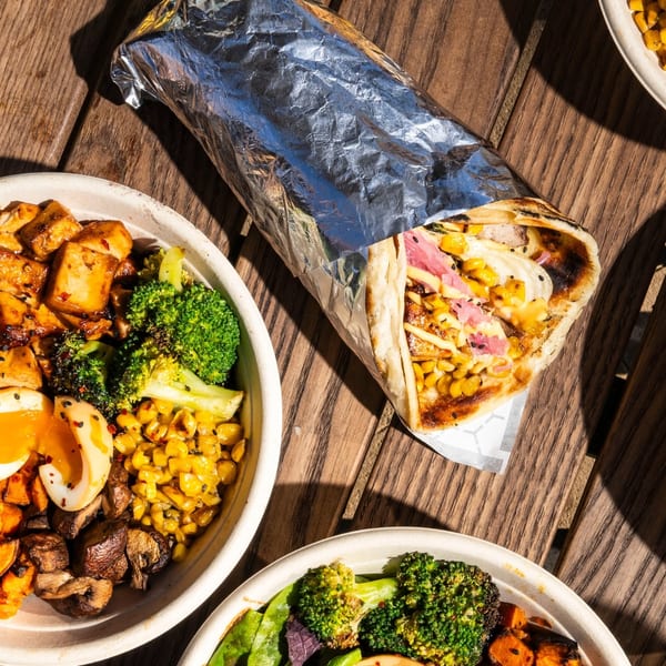 A large burrito and two vibrant bowls with eggs, broccoli, corn, and protein on a wooden table.