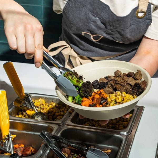 Chef adds broccoli to a custom bowl with meat, corn, and sweet potatoes at a serving station.