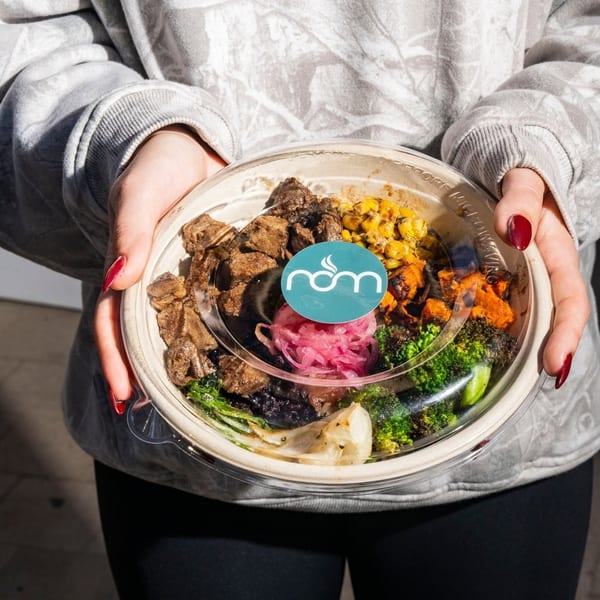 Hands holding a nom meal bowl with beef, corn, sweet potatoes, broccoli, and pickled onions.