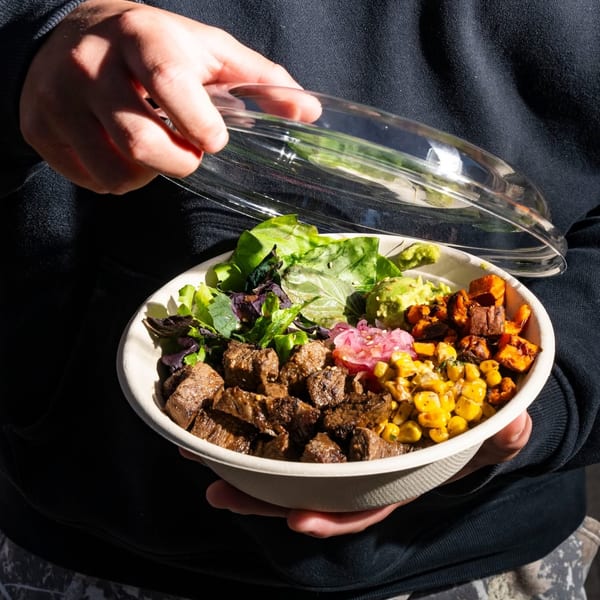 Hands holding a takeout bowl filled with steak, guacamole, corn, sweet potatoes, and greens.