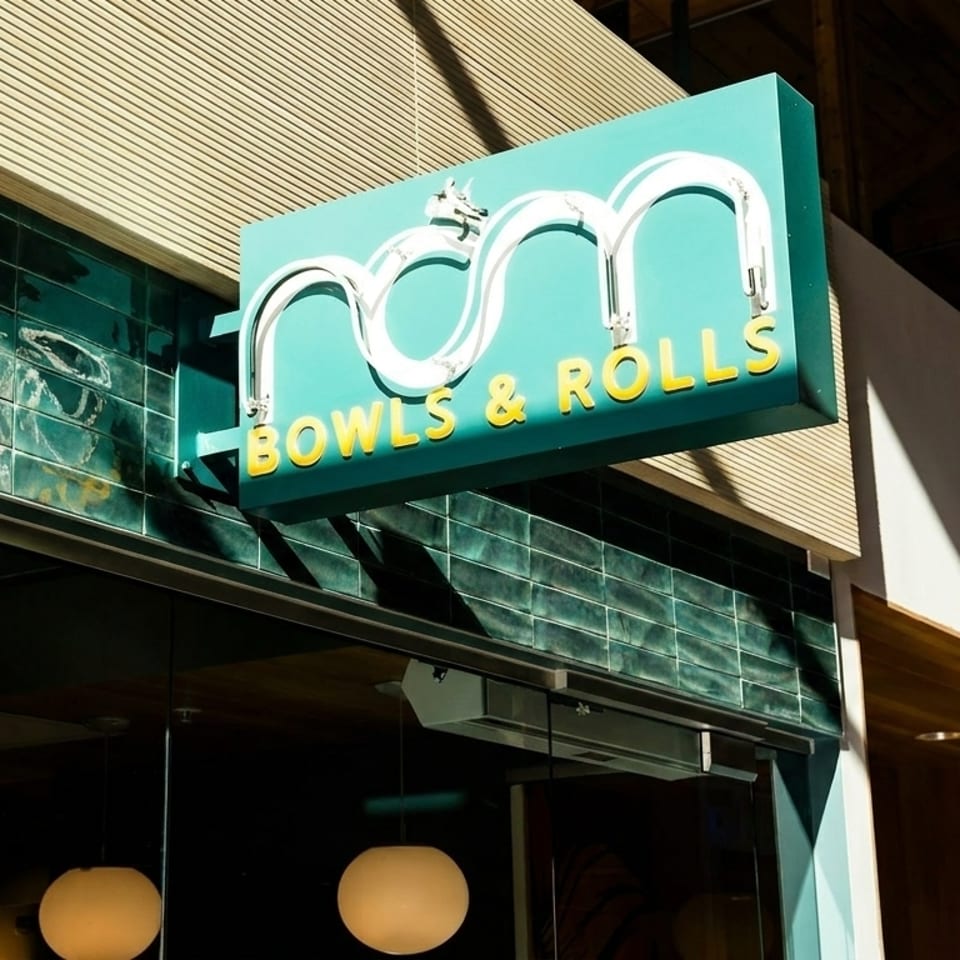 A teal "Bowls & Rolls" sign with white neon letters hangs above a restaurant entrance.
