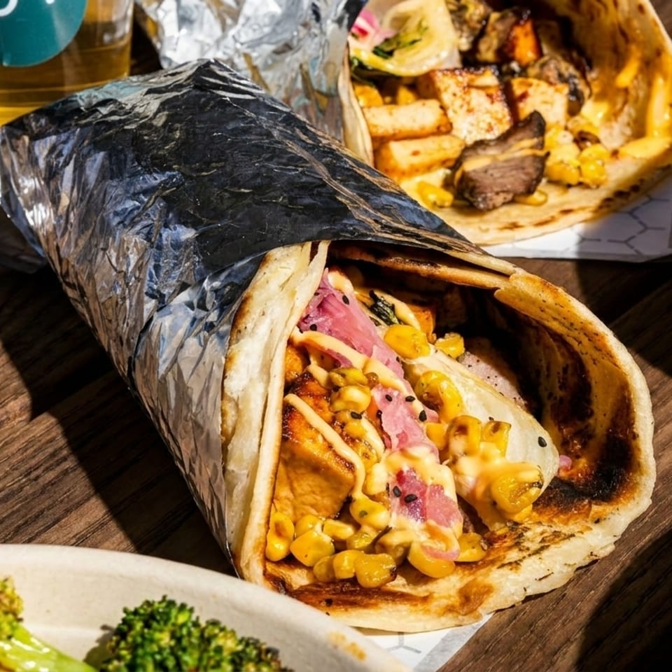 A close-up of a foil-wrapped burrito with corn, meat, and sauce, surrounded by other dishes.