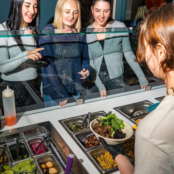 Three young women watch a server prepare a custom food bowl at a restaurant counter.
