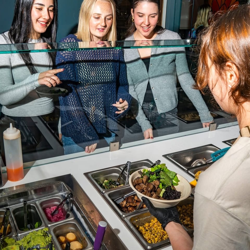 Three young women watch a server prepare a custom food bowl at a restaurant counter.